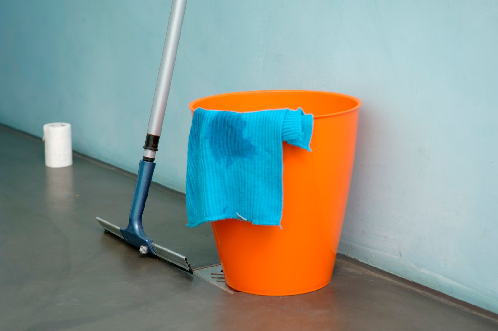 An orange bucket, a blue cloth and a squeegee against a pale blue wall