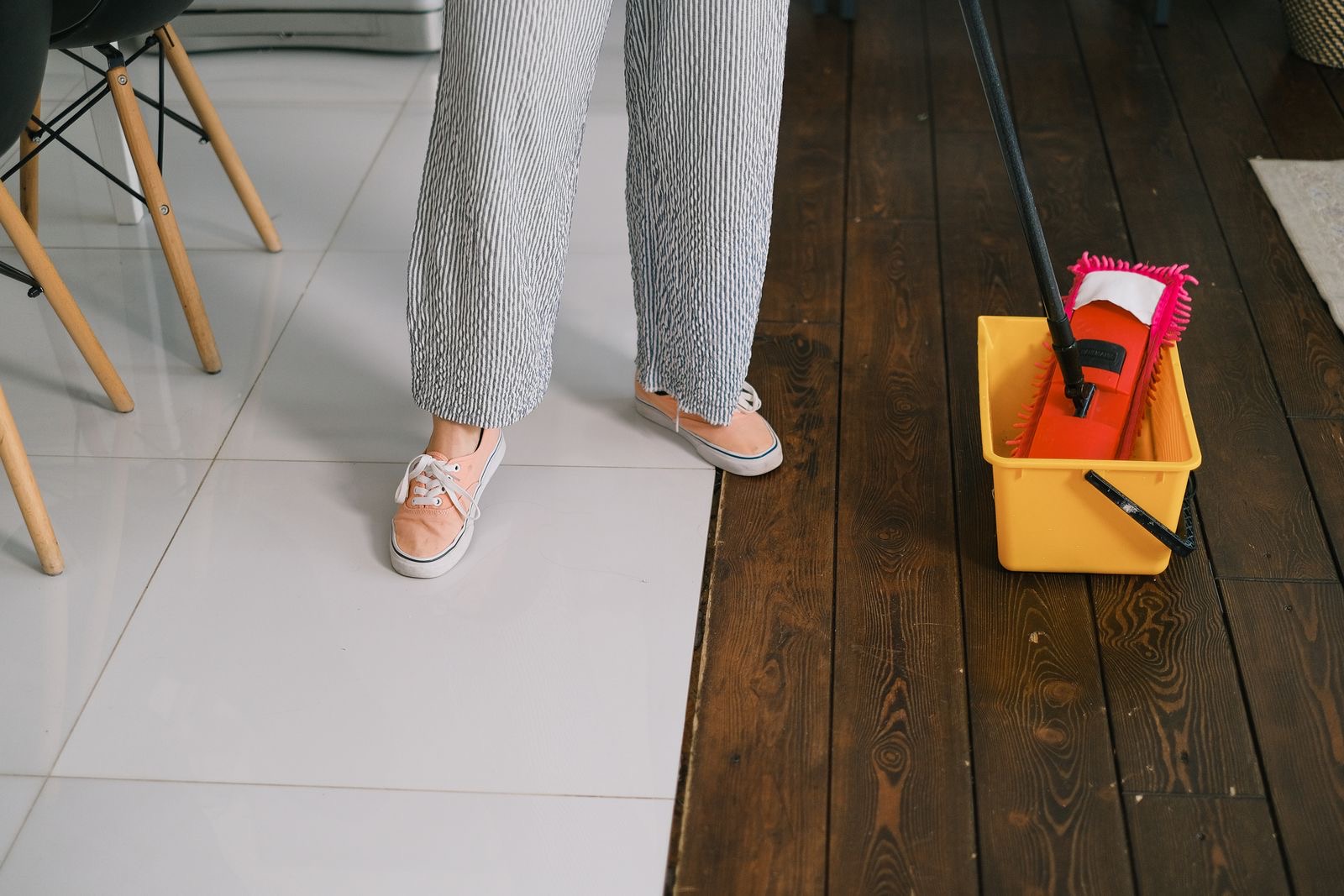 Feet beside a yellow mop bucket and a flat mop, where kitchen tiles meet floorboards